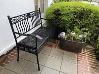 Black painted metal outdoor bench positioned next to metal tub planter with solar lights in an outdoor patio area.