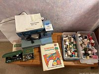 Full lot showing Riccar 3300 sewing machine, instruction manual, Singer sewing book, trays with thread spools, Singer automatic attachment box with attachments, and booklet on wood table against a textured wall.