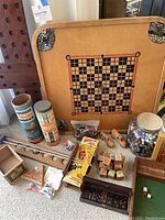 Overview photo showing vintage Carrom board, Tinkertoys tins, plastic bricks tube, large marble jar, and wooden toys and games spread out on floor.