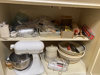 Shelf with white Sunbeam hand mixer, ceramic canister, nested ceramic bowls, and various baking accessories in boxes and bags