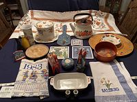 Full lot overview showing all items including tablecloth, teapot, canister, plates, towels, trivets, plaques, figurines and decorative items arranged on a table.