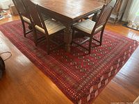 Vintage red patterned rug under dining table with chairs on a wooden floor. The rug's pattern includes intricate floral and geometric designs in red, black, cream, and tan colors.