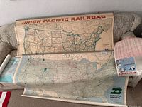Two large vintage USA rail maps rolled out on couch showing state borders and railway routes, with visible aging and creasing.