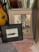 Two framed original signed watercolor paintings shown leaning against a wall near a guitar. One larger piece with a wide silver frame and an abstract texture composition. One smaller piece in a simple black frame with a winter landscape.