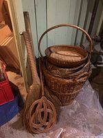 Photo showing 5 wicker baskets stacked and two straw brooms leaning beside them, with one broom having tightly bound straw bristles and a woven wooden tool also visible in front.
