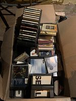 Box filled with stacked CDs and cassette cases, showing variety of albums and some visible labels arranged in rows within a single cardboard box.