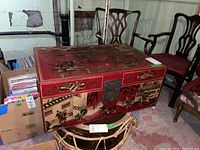 Front view of the rectangular trunk showing the latch, painted figures in garden scenes, red base color, and some wear.