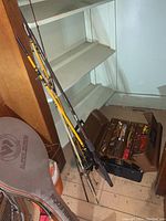 Photo showing five fishing rods leaning against a wall next to the tackle box on a wooden floor.