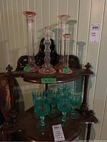 Four pink glass candle holders arranged on an antique two-tier wooden shelf, with eight green glass stemmed glasses positioned below them on the lower tier, showing overall lot composition and scale.