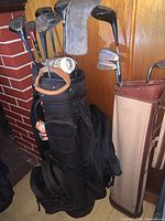 Photo showing two golf bags with assorted golf clubs, one black soft-sided, one vintage-style beige and brown, with various clubs including drivers with covers and irons.