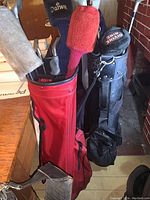 Photo showing red and black golf bags with clubs and head covers on wooden floor near brick wall.