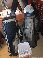 Two golf bags standing side by side; one black and beige with clubs, one olive green with clubs; white pail of golf balls in front.