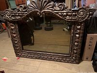 Full view of the wood framed mirror leaning against a surface showing detailed carved patterns around frame edges and crest on top center