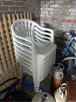 Stack of seven white plastic lawn chairs in a garage setting against a brick wall, showing dirt and usage marks on surfaces