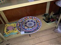 Three decorative ceramic platters on a shelf: large round platter with colorful circles, rectangular yellow platter with dog design, and ceramic basket bowl with handles.