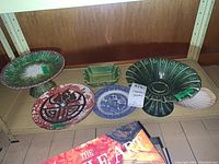 Shelf displaying multiple serving pieces including green pedestal cake stand, various platters, plates, and trivet.