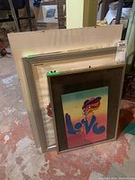 Three posters standing vertically on floor; two framed and one laminated between glass and board. The closest framed poster features colorful psychedelic 'Love' text.