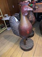 Front and side views of large wooden carved rooster statue showing red brown paint and worn surface.