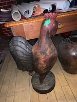 Front and side view of a large rustic wooden rooster with carved details and painted features including red comb and earthy body colors.