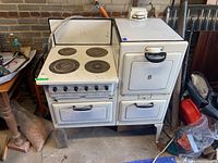 Overall front view of the vintage gas stove showing four circular burner plates, three ovens with metal handles, and white enamel finish with some visible wear.