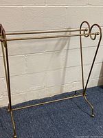 Gold-colored metal quilt rack against a beige brick wall, showing full front and side profile including the decorative swirl side supports and dual horizontal bars for holding quilts or blankets.