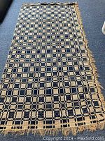 Top view of one piece of antique fabric coverlet showing blue and cream geometric patterned textile with frayed edges on a dark carpet.