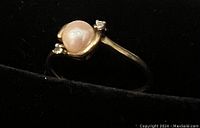 Ring set in 10k yellow gold with solitaire pearl and two small clear stones, shown upright in black background.