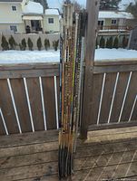 Photo of nine wooden hockey sticks standing against wood fence with snowy background.