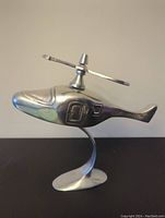 Side profile of aluminum helicopter sculpture with rotor blades attached on top and curved boat-shaped base.