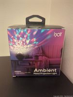 Front of box showing iJoy Ambient Mood Projection Light projecting colorful light patterns in a living room setting. Text indicates contents include lamp with USB cord and user manual.