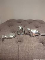 Three small polished aluminum animal sculptures on a gray tufted fabric surface: a loon, frog, and beaver, showing smooth, reflective forms.