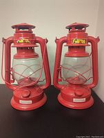 Two red kerosene lanterns side by side showing front and labels.