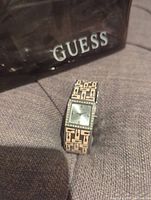 Photo of the GUESS watch with box partially visible, showing the pink and black patterned band and silver square face with rhinestone bezel.