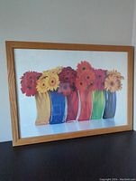 Front view of framed print featuring colorful gerbera daisies in multicolored glass vases, wooden frame, and clear white background.