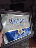 Front view of the rectangular mirrored Corona Extra sign held by hand indoors, showing blue and gold text and graphic details with clear reflections.