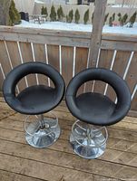 Pair of two black faux leather bar stools with circular backs and chrome pedestal bases shown outdoors by wooden fence. Both stools in lightly used condition with visible wear on seats.