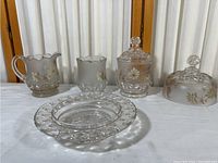 Five-piece set of crystal glass items featuring frosted and clear glass with gold-tone floral etching. Items include two pitchers or creamers, two lidded jars, and one round dish.
