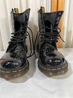 Front view of black patent leather Doc Martens boots with yellow stitching and lace-up design.