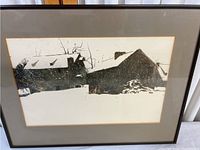 Framed monochrome print of a snowy cabin with visible snow on roof and trees, displayed against a neutral-colored matting and enclosed in a black frame under clear glass.