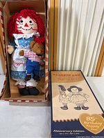 Raggedy Ann and Raggedy Bear dolls lying in original box, box lid shown with 85th birthday anniversary labeling.