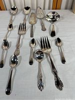 Collection of 11 silver plated silverware items including spoons, forks, and pie server laid out on white cloth showing varied sizes and ornate designs.