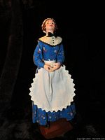 Full front view of handmade Simpich Mrs. Cratchit doll in blue floral dress, white apron and cap, holding a small bowl, standing on a wooden base.