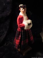 Front and side views of a Simpich wooden lady caroler doll from 1986 wearing a red jacket, red and black plaid skirt, white fur muff with holly, and a black hat with a red ribbon, mounted on a wooden base.