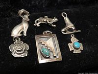 Photo of six sterling silver charms laid out on a black surface. Charms include a cat, frog, ice skate, rose, rectangular charm with turquoise, and a bird charm with turquoise.
