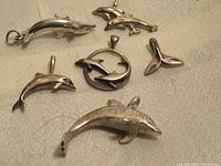 Six sterling silver dolphin-themed charms laid out on fabric background. Includes single dolphin pendants, two dolphins inside a circle, and a dolphin tail.