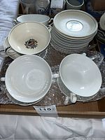 Box containing stacked white plates with central floral basket pattern and radial embossed rim detail, surrounded by bowls including some with floral motifs and handles.