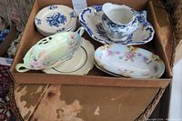 Photo of assorted decorative plates, floral porcelain serving trays, and Royal Staffordshire blue and white pitcher with tray inside a cardboard box