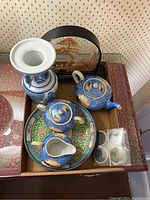 Photo showing blue and white porcelain vase, blue and peach motif porcelain teapot, creamer, sugar bowl, floral plate, and glass napkin rings.
