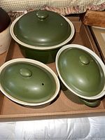 Three green ceramic lidded pots arranged in a box. The pots have a round shape with lids featuring knob handles and vent holes. The largest pot is visible at the back, with two smaller pots in front at angles.