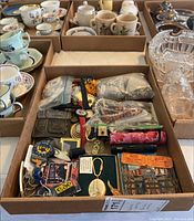View of assorted costume jewelry, beads in bags, watches, keychains, belt buckles, collectible pins, cards, and stamps all contained in a cardboard box with other boxed dishware visible in background.